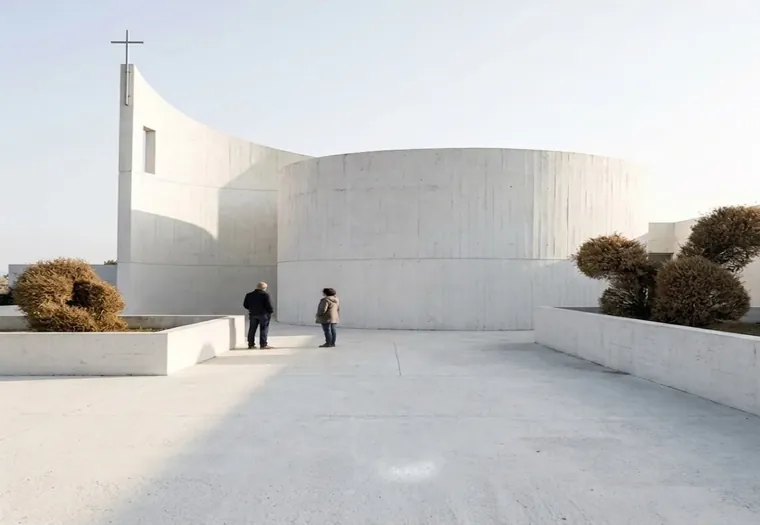 Render: a white concrete chapel with a curved wall and a slender tower, two people on a bare forecourt