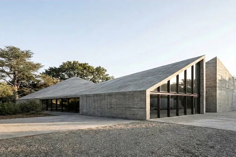 Render: a concrete building under a folded roof rising to a tall glazed gable, gravel forecourt